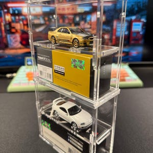 May include: Two miniature toy cars, a gold Nissan Skyline GT-R R34 and a white Toyota Supra, displayed in a clear acrylic display case. The cars are on top of their original packaging boxes.