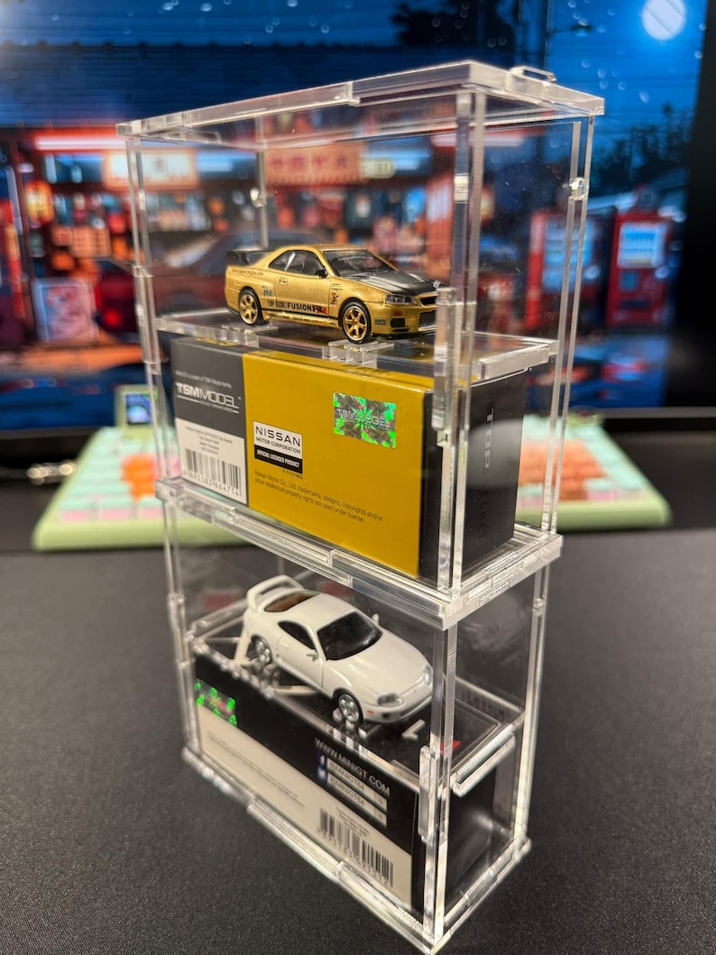 May include: Two miniature toy cars, a gold Nissan Skyline GT-R R34 and a white Toyota Supra, displayed in a clear acrylic display case. The cars are on top of their original packaging boxes.