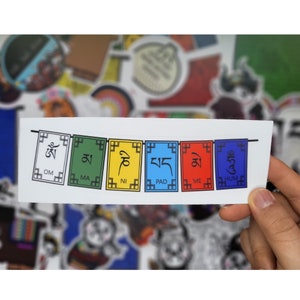 May include: A sticker featuring a string of colorful prayer flags. Each flag is a different color: white, green, yellow, blue, red, and blue. The flags have black symbols and text. The sticker is held by a hand.