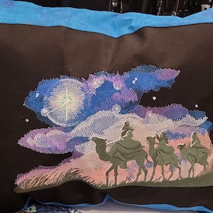 May include: Black pillow with an embroidered scene of three figures riding camels under a starry sky. The embroidery features a bright star, clouds in shades of blue and purple, and a blue border.