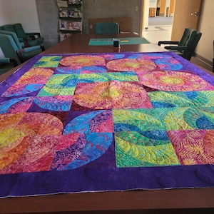 May include: A large, rectangular quilt featuring a vibrant, multi-colored pattern of swirling circular and leaf-like motifs. The fabric displays a spectrum of bright pink, orange, yellow, green, and blue hues, all bordered by a solid purple fabric.