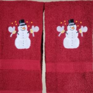 May include: Two red hand towels with an embroidered snowman design. The snowman has a black top hat, a carrot nose, and is holding mittens. The towels have a solid red border.