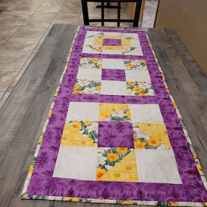 May include: Purple and white table runner with a yellow floral pattern. The runner is quilted and has a border of purple fabric with a floral design.