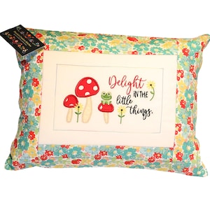 May include: A white pillow with a floral print border. The center of the pillow has a white background with embroidered red mushrooms, a green frog, and the text "Delight in the little things."