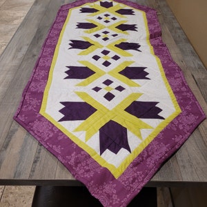 May include: A quilted table runner with a white center, featuring a geometric pattern in purple and yellow. The border is a floral print in shades of purple. The runner is rectangular and designed to decorate a table.