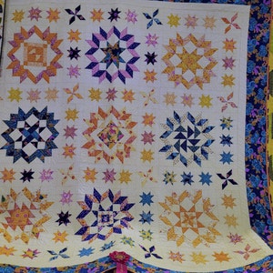 May include: A quilt with a white background and a variety of colorful star and geometric patterns. The quilt is edged with a blue and purple floral border. A blue ribbon with a gold seal is attached to the bottom of the quilt.