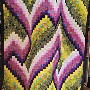 May include: A quilt with a geometric pattern of squares in shades of yellow, green, purple, and white. The squares are arranged in a swirling pattern that creates the illusion of movement.