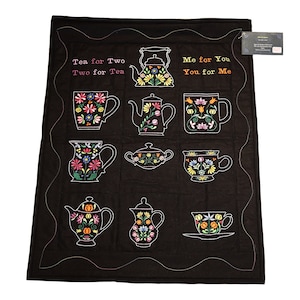 May include: Black quilted wall hanging with a white embroidered design of teapots, cups, and saucers. The text "Tea for Two Two for Tea" and "Me for You You for Me" is embroidered on the wall hanging.