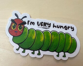 May include: A die-cut sticker featuring a cartoon caterpillar with a red head, yellow eyes, and a segmented green body. The caterpillar has a grumpy expression with thick eyebrows and the text "I'm VERY hungry" written in black.
