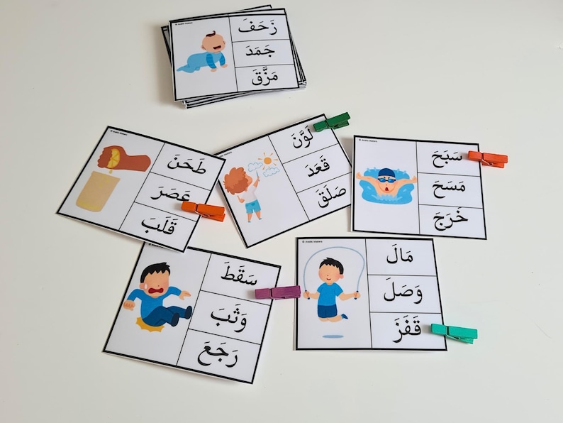May include: Educational flashcards with Arabic words and illustrations. Each card features a picture and corresponding word, secured with colorful clothespins. The cards depict various actions and objects, ideal for early learning.