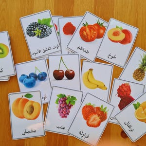Puede incluir: Un conjunto de tarjetas didácticas de frutas, cada una con una colorida ilustración de diferentes frutas, como moras, tomates, melocotones, arándanos, cerezas, plátanos y fresas. Las tarjetas están sobre una superficie de madera.