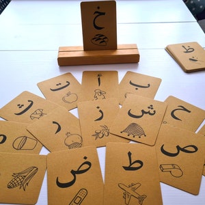 May include: A set of flashcards with Arabic letters and illustrations. The cards are brown with black lettering and drawings of objects like an apple, a pomegranate, and a whistle. A wooden card holder displays a card. The cards are arranged on a white surface.