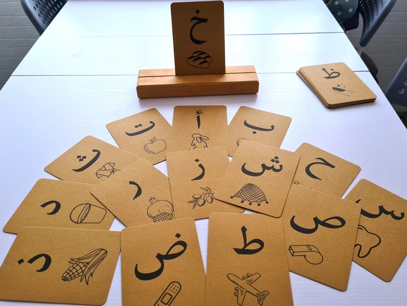May include: A set of flashcards with Arabic letters and illustrations. The cards are brown with black lettering and drawings of objects like an apple, a pomegranate, and a whistle. A wooden card holder displays a card. The cards are arranged on a white surface.