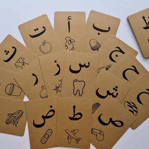 May include: A collection of flashcards with Arabic letters and corresponding illustrations in black ink on a brown cardstock. The images include an apple, a rabbit, an acorn, a tooth, and a bandage.