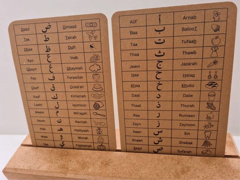 May include: Two rectangular flashcards with Arabic letters and corresponding English transliterations and images. The cards are brown with black text and illustrations. The cards are displayed on a wooden stand.