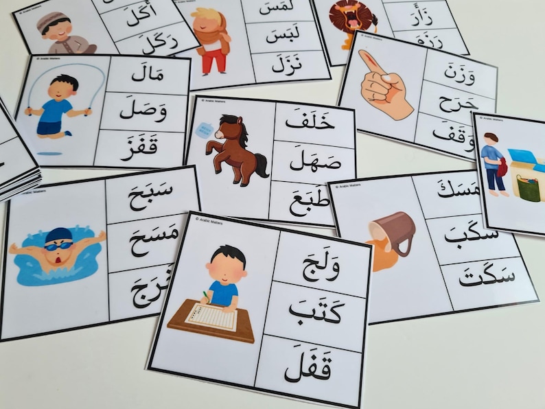 May include: A collection of flashcards with Arabic words and illustrations. Each card features a word in Arabic script paired with a corresponding image, such as a horse, a person swimming, or a person writing. The cards are white with black text and colorful illustrations.