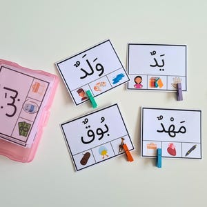 Peut inclure: Un étui en plastique rose contenant des cartes flash avec du texte et des illustrations en arabe. Quatre cartes flash individuelles avec des mots arabes et des images correspondantes sont affichées. Des pinces à linge colorées sont éparpillées autour des cartes.