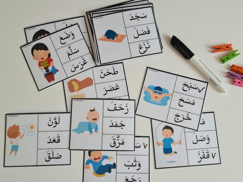May include: A collection of flashcards with Arabic text and illustrations of children performing various actions. A black marker and colorful clothespins are also visible. The cards are designed for educational purposes.