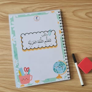May include: A spiral-bound notebook with a cover featuring Arabic text and illustrations of a globe, scissors, and a pencil cup. A black marker and a red eraser are next to the notebook on a wooden surface.