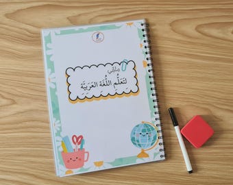 May include: A spiral-bound notebook with a cover featuring Arabic text and illustrations of a globe, scissors, and a pencil cup. A black marker and a red eraser are next to the notebook on a wooden surface.