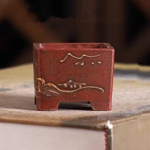 May include: A small, square, red ceramic bonsai pot with a gold design. The pot has a slightly raised rim and four small feet.