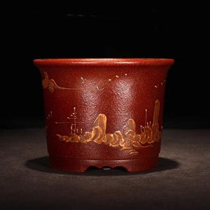 Puede incluir: Una maceta de arcilla roja con un diseño dorado de un paisaje con montañas y árboles. La maceta tiene una forma redondeada y una abertura ancha.