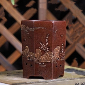 Puede incluir: Una maceta hexagonal de terracota con un esmalte marrón y detalles pintados en oro. La maceta presenta una escena de paisaje con montañas, árboles y pájaros.