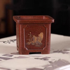 Puede incluir: Un macetero cuadrado de terracota con un esmalte marrón y detalles pintados en oro. El macetero tiene un borde elevado y una base cuadrada.