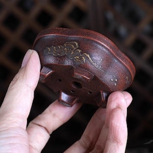 Puede incluir: Una maceta de bonsái de terracota con un esmalte marrón y un diseño de hoja de oro. La maceta tiene cuatro patas y un agujero de drenaje en la parte inferior.