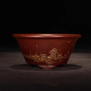 May include: A red clay bonsai pot with a gold painted landscape design. The pot has three small feet.