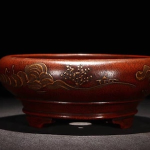 May include: A red ceramic bonsai pot with a gold design of clouds and a tree. The pot has three legs.