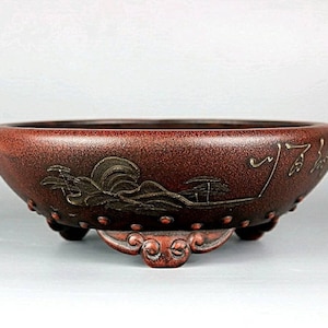 May include: A red clay bonsai pot with a gold and silver design of a wave and a mountain. The pot has three feet and is decorated with small dots.
