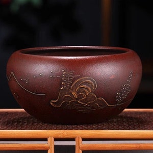 Puede incluir: Un cuenco de té de cerámica rojo oscuro con un diseño dorado de montañas y árboles. El cuenco está sentado en un soporte de madera.
