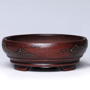 Może przedstawiać: Czerwona ceramiczna doniczka do bonsai z trzema nóżkami i złotym wzorem gór i ptaków.
