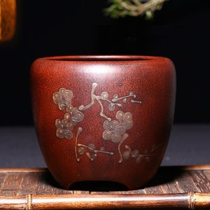 May include: A dark red ceramic pot with a wide mouth and a rounded base. The pot has a decorative design of gold-colored flowers and branches.