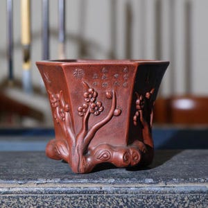 Può includere: Un vaso in terracotta rossa a forma esagonale con un designo scolpito di un albero con fiori. Il vaso ha una base rialzata con tre gambe.
