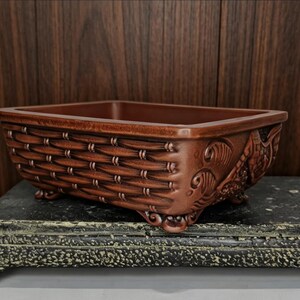 May include: A rectangular, brown ceramic planter with a woven basket design on the sides. The planter has decorative relief carvings on the ends and sits on small feet. The interior is a darker shade of brown.