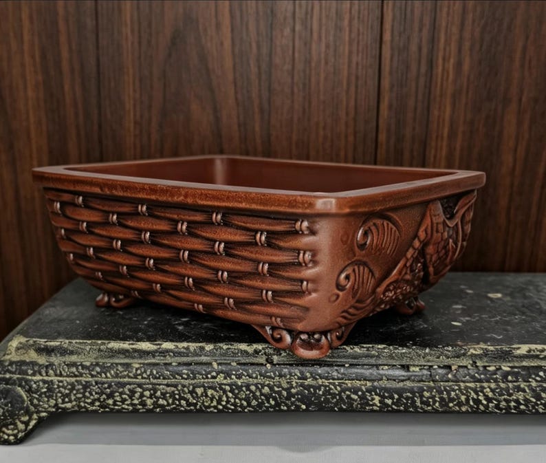 May include: A rectangular, brown ceramic planter with a woven basket design on the sides. The planter has decorative relief carvings on the ends and sits on small feet. The interior is a darker shade of brown.