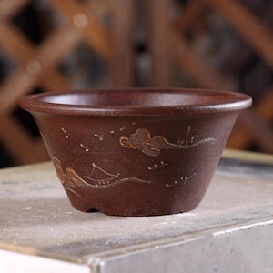 Puede incluir: Una maceta de bonsái de cerámica marrón con un diseño ancho y poco profundo. La maceta presenta ilustraciones de paisajes grabadas con un barco y nubes. La maceta está sobre una superficie de color claro.