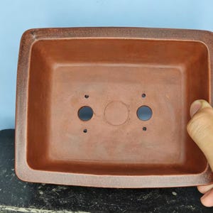 May include: A rectangular, reddish-brown bonsai pot with rounded corners. The pot has two drainage holes and a central hole. The pot is made of ceramic and is designed for growing bonsai trees. The pot is held by a hand.