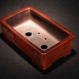 May include: A rectangular red bonsai pot with a dark brown interior and three drainage holes. The pot has a raised rim and a decorative design on the side.