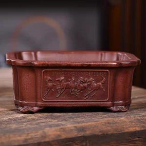 Peut inclure: Un pot carré en terre cuite avec un bord surélevé et quatre pieds. Le pot présente un motif de chevaux courant en cercle sur le devant.