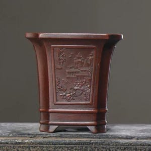 Könnte beinhalten: Ein rechteckiger Keramik-Blumentopf in Dunkelrot mit einem erhabenen Design eines Gebäudes und Bäumen. Der Topf hat einen quadratischen Boden und vier Beine.
