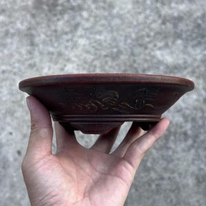May include: A small, round, reddish-brown bonsai pot with a decorative relief design around the rim. The pot has a low profile and is supported by small feet. The relief design appears to depict a landscape scene.