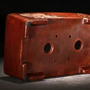 May include: A terracotta bonsai pot with four drainage holes on the bottom. The pot has a textured surface and a reddish-brown color.