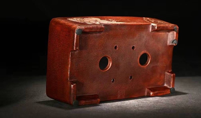 May include: A terracotta bonsai pot with four drainage holes on the bottom. The pot has a textured surface and a reddish-brown color.
