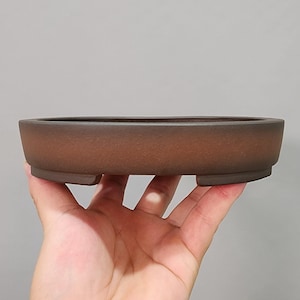 Könnte beinhalten: Ein brauner, ovaler Bonsai-Topf mit niedrigem Profil. Der Topf hat eine leicht strukturierte Oberfläche und wird von vier kleinen Füßen getragen. Der Topf wird in einer Hand vor einem neutralen Hintergrund gehalten.