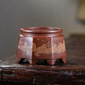 Puede incluir: Una pequeña maceta de cerámica de color marrón rojizo con forma hexagonal y tres patas pequeñas. La maceta presenta un diseño de paisaje con montañas y árboles en un color más claro en los laterales. La maceta está sobre una superficie de madera oscura.