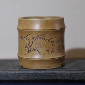 Peut inclure: Un pot en céramique brun avec un motif de bambou et de la calligraphie chinoise. Le pot a une surface texturée et une forme légèrement arrondie.