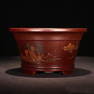 May include: A red ceramic bonsai pot with a gold painted landscape design. The pot has a rounded shape and three short legs.